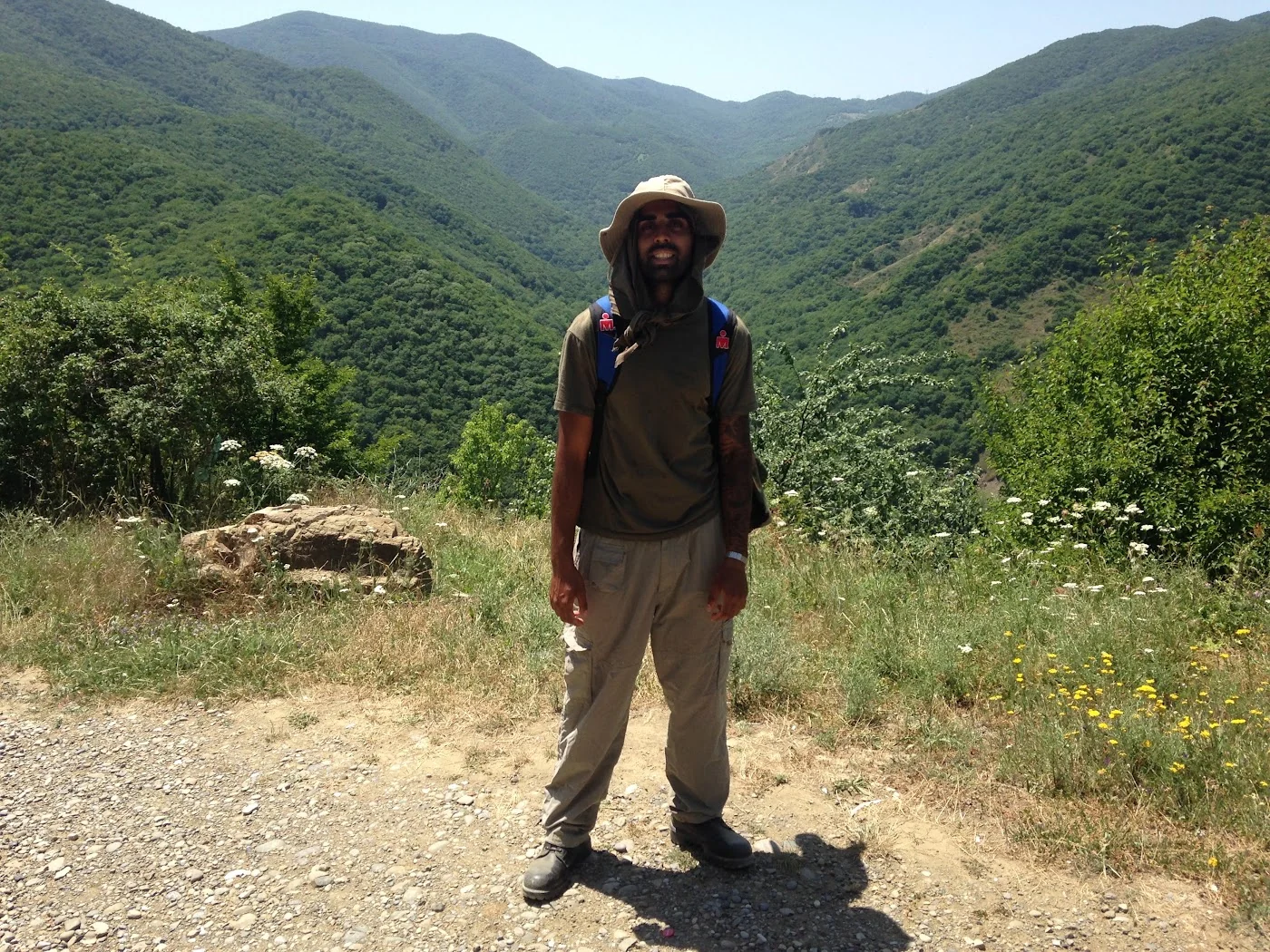 Deepak Shukla standing with a backpack and sun hat on gravel above a wooded valley