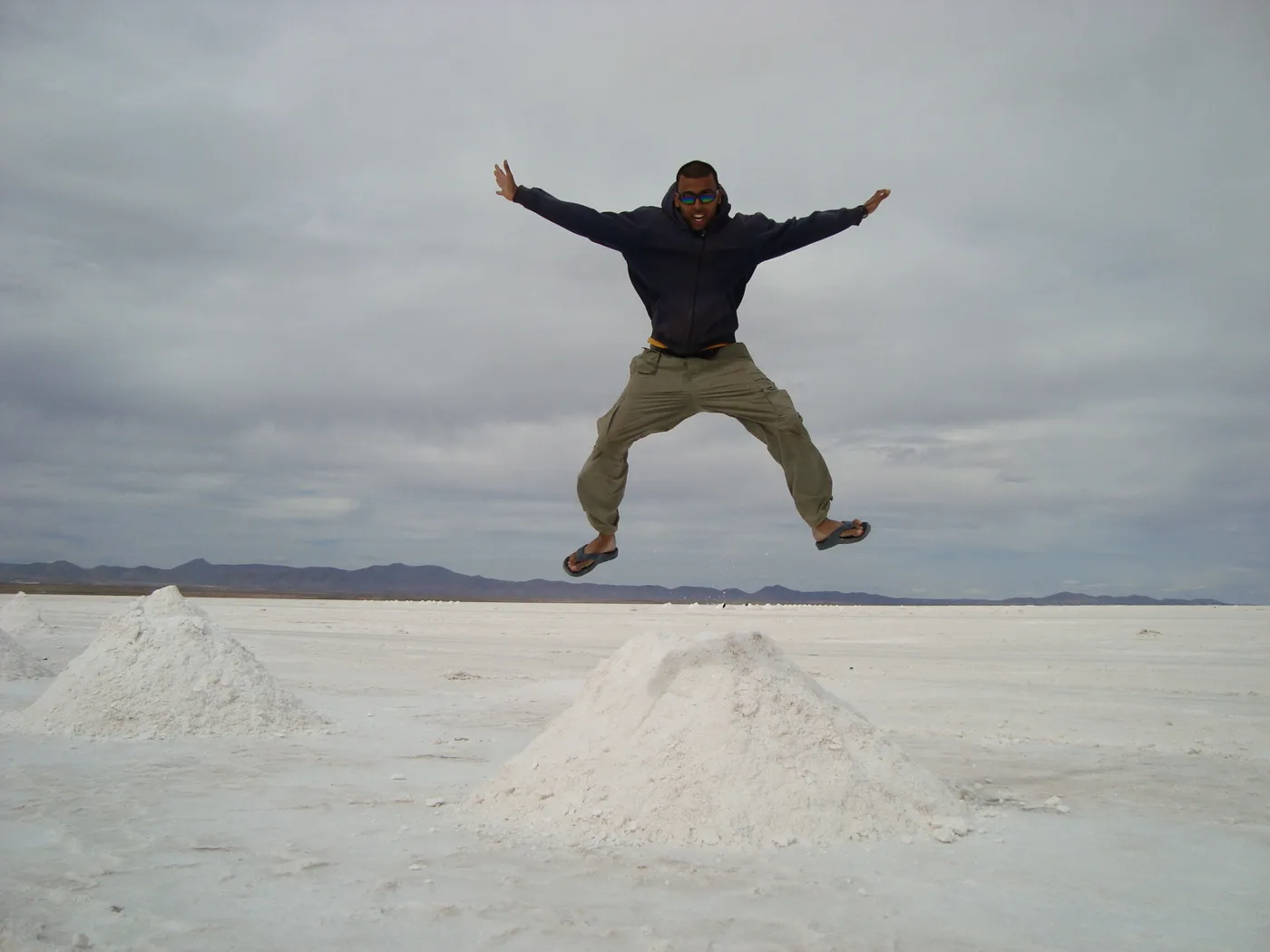 Deepak jumping with arms spread above the pale Uyuni salt flats beneath a cloudy sky
