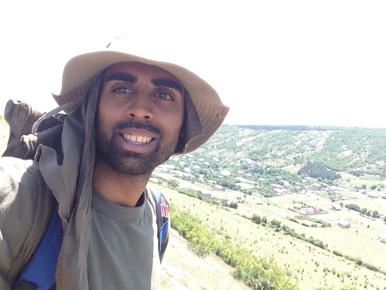 Deepak Shukla wearing a sun hat and backpack above a green valley