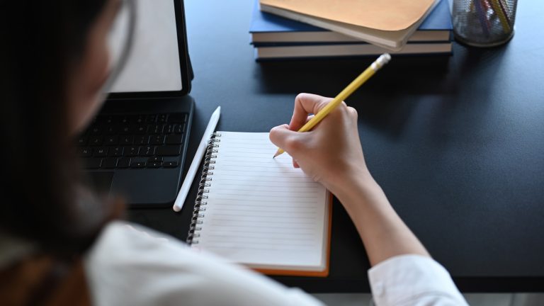 Close up view of young businesswoman using computer tablet and writing on empty notebook.